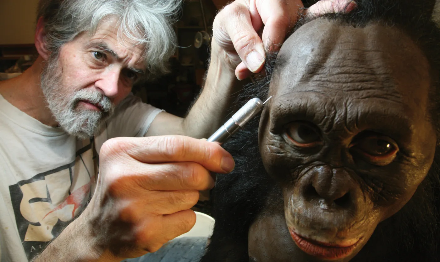 John Gurche in his studio, sculpting the face of a hominin reconstruction with a fine modeling tool.